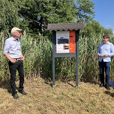 Zwei Männer stehen neben einer Infotafel am Wegesrand.