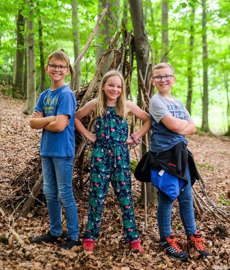 Drei Kinder vor selbstgebautem Unterschlupf im Wald