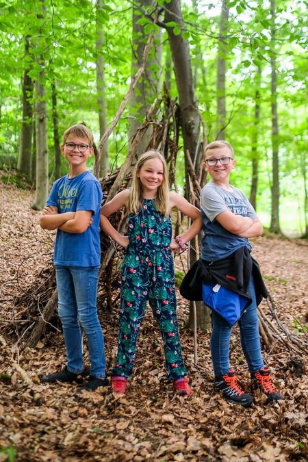 Drei Kinder vor selbstgebautem Unterschlupf im Wald