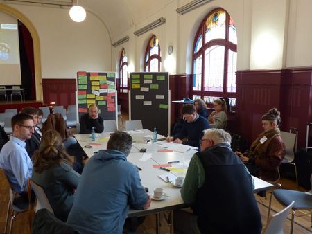 Eine Gruppe Menschen sitzt um einen Tisch und hält einen Workshop, im Hintergrund ein Board mit Postits