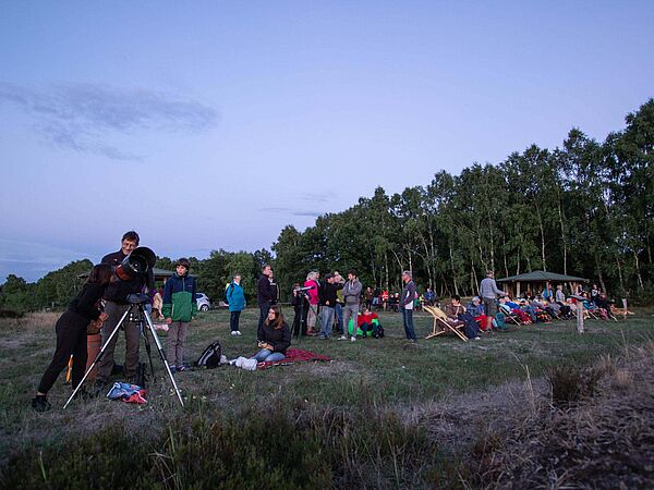 Auf einem Hügel treffen sich etwa 40 Menschen um in der Abenddämmerungen Sterne zu beobachten. Es wurde ein großes Teleskop aufgebaut.