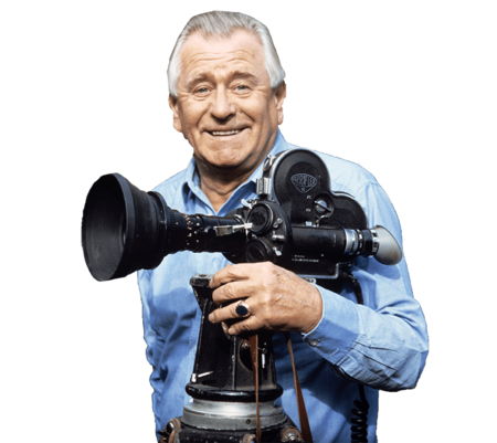 Heinz Sielmann hält in einer Hand eine große schwarze Filmkamera aus den 70er Jahren. Er lächelt den Fotografen an und trägt ein hell blaues Hemd. 