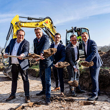 Männer im Anzug heben Spaten, auf denen Erde ist, hinter ihnen steht ein Bagger