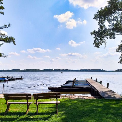 Vom Ufer des Groß Schauener Sees blickt man über eine Holzbank hinweg auf einen kleinen hölzernen Steg an dem mehrere Ruderboote festgemacht sind. Am Himmel über dem blauen See sind wenige weiße Wolken zu sehen, die Sonne spiegelt sich im Wasser.