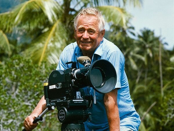 Sielmann beim Filmdreh unter Palmen Heinz Sielmann steht lächelnd in einem kurzärmeligen Hemd an der Kamera, hinter ihm eine tropische Landschaft