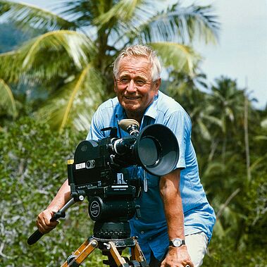 Sielmann beim Filmdreh unter Palmen Heinz Sielmann steht lächelnd in einem kurzärmeligen Hemd an der Kamera, hinter ihm eine tropische Landschaft