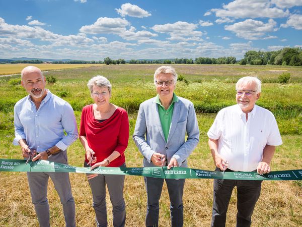 Vier Personen - eine Frau und drei Männder - halten ein grünes Band in der Hand und sind kurz davor, dieses mit Scheren zu zerschneiden. Im Hintergrund leuchtet im Sonnenlicht die Biotopfläche.