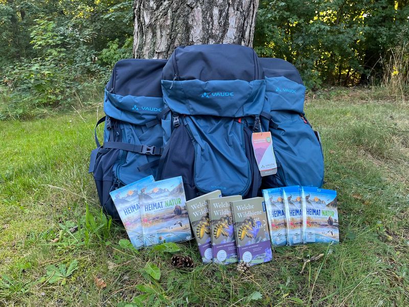 Drei blaue Rucksäcke von VAUDE an einen Baum gelehnt, davor stehen DVDs und BlueRays von Heimat Natur und Bücher mit Wildbienenführer