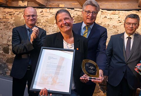 Preisträgerin Agnes Becker hält Ihre Urkunde und den Preis in den Händen und lächelt in die Kamera. Im Hintergrund stehen Fritz Brickwedde, Stiftungsratsvorsitzender und zwei weitere Herren.