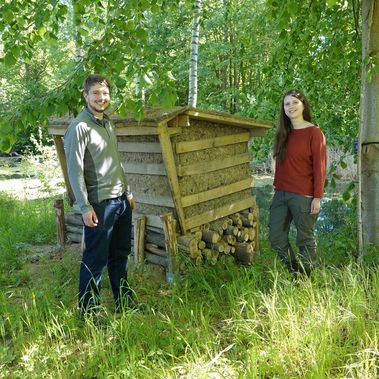 Junger Mann und Frau stehen von zwei Seiten neben einer Nisthilfe aus Holz