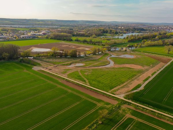 Aus der Vogelperspektive sind die neuen Strukturen auf den Flächen zu erkennen. Dazu zählt ein Weiher und mehrere temporäre Tümpel. Links und rechts sind intensiv genutzte Ackerflächen. Am Horizont befinden sich einige Gebäude der Stadt Göttingen. Die Leine fließt durch dieses Areal.