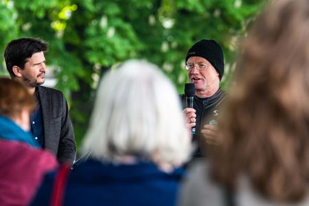 Ein Mann mit Mütze spricht in ein Mikrofon, vor und neben ihm stehen Menschen
