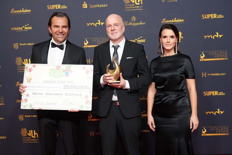 Robert Dahl, Peter Nitschke und Yara Hoffmann stehen vor einer Fotowand mit Trophäe und großem Spendenscheck in der Hand.