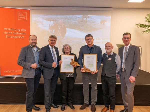 Preisverleihung Ehrenpreis Mehrere Personen vor einer Bühne, zwei halten Urkunden in die Kamera