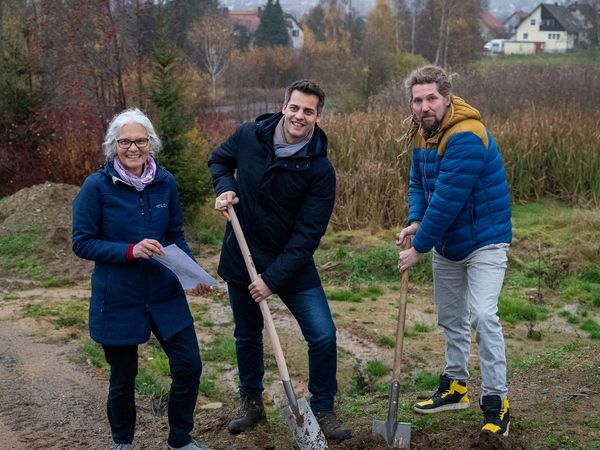 Drei Personen stehen im Frühwinter auf einer Brache. Links steht eine Frau mit dickem blauen Mantel und grauen Haaren und hält ein Dokument in den Händen. In der Mitte hält ein Mann im Wintermantel einen Spaten auf den er einen Fuß gestellt hat. Rechts steht ein weiterer Mann in einer Daunenjacke und hält ebenfalls einen Spaten.