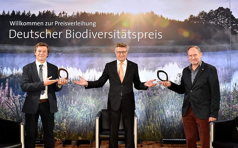 3 Herren stehen auf einer Bühne und halten einen Preis in der Hand bzw. bekommen diesen vom Mann in der Mitte überreicht