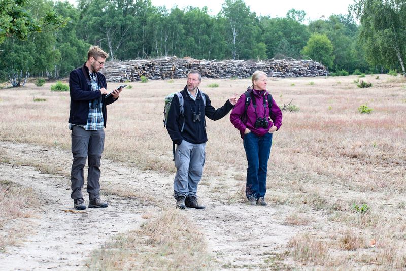 Drei Menschen stehen in einer sandigen Graslandschaft und hören, ob Vögel singen. Der junge Mann ganz links hält ein Tablett, ein älterer Mann in der Mitte zeigt nach rechts, eine Frau steht ganz rechts und blickt in die Landschaft. Alle tragen Outdoor-Kleidung.